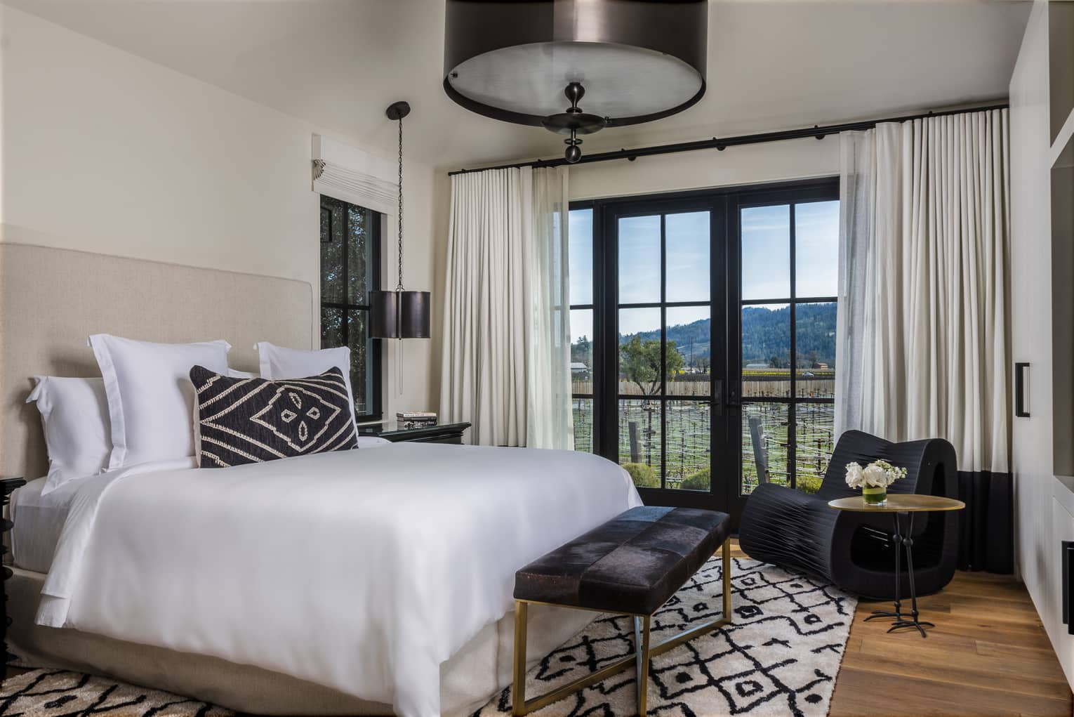 Luxury resort bedroom with upholstered bed, patterned pillow, black bench, modern black chair and large windows with vineyard and hillside views