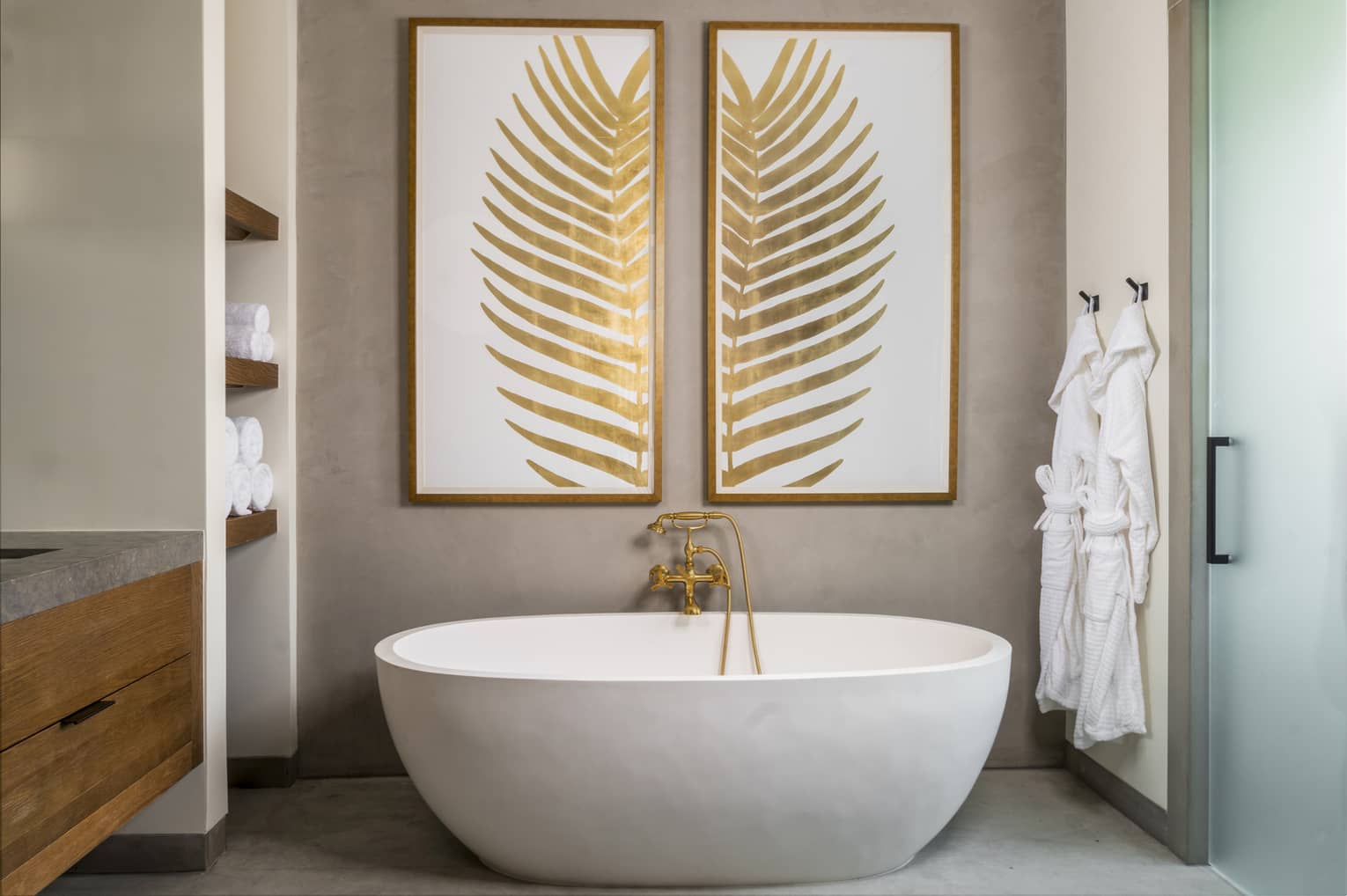 Resort bathroom with a freestanding oval bathtub, gold faucet, two pieces of leaf artwork, built-in towel shelves and two white robes hanging by frosted door