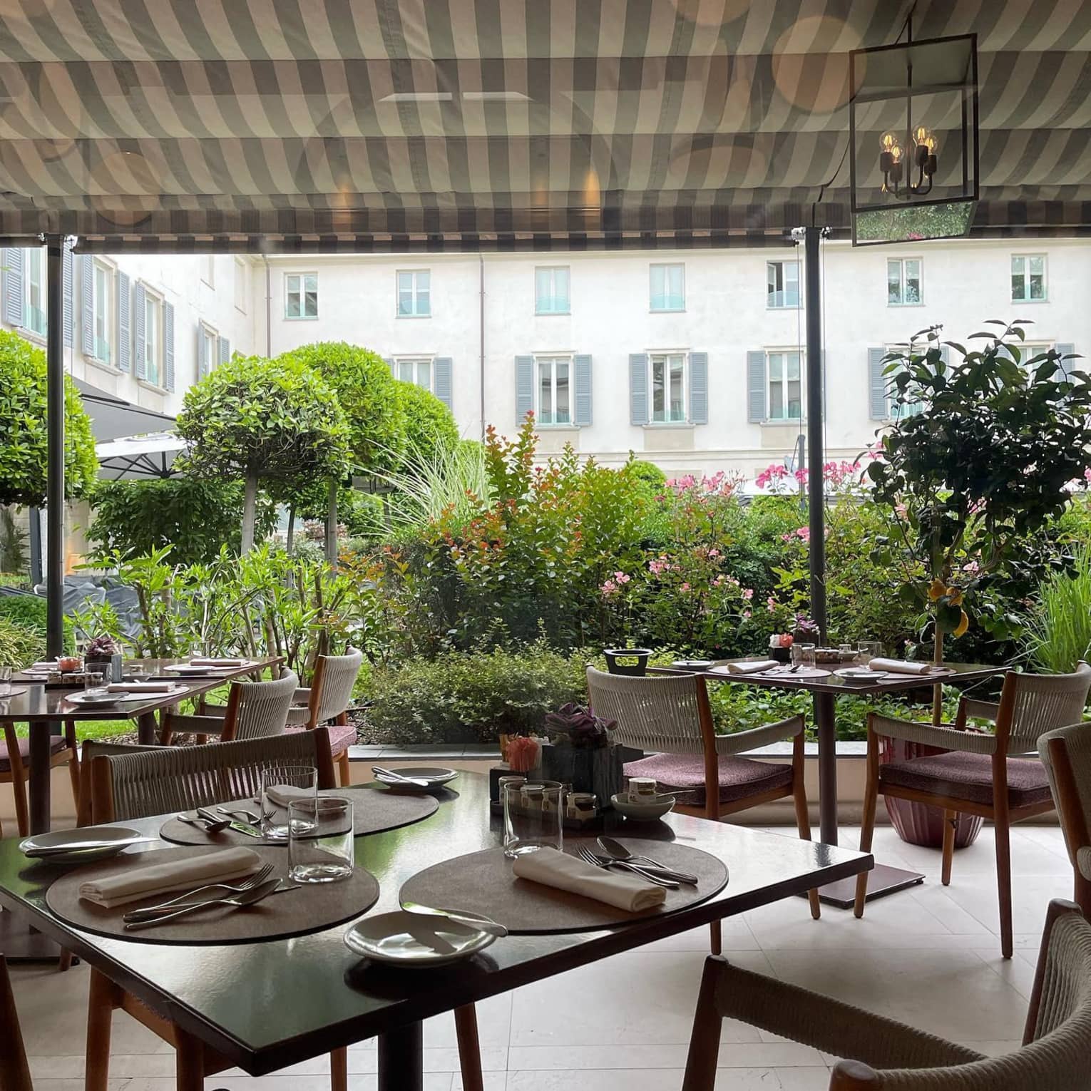 Several dining tables are set with silverware and glasses underneath a fabric covering on an outdoor terrace surrounded by greenery, at the Four Seasons Hotel Milan