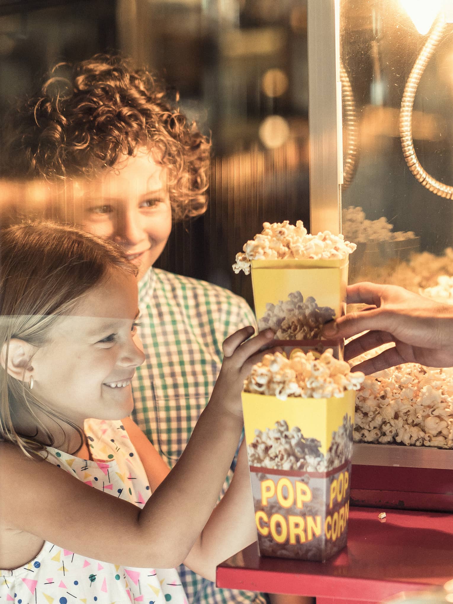Two children smile in the warm glow of a movie-style popcorn machine as they receive boxes of popcorn from a vendor.
