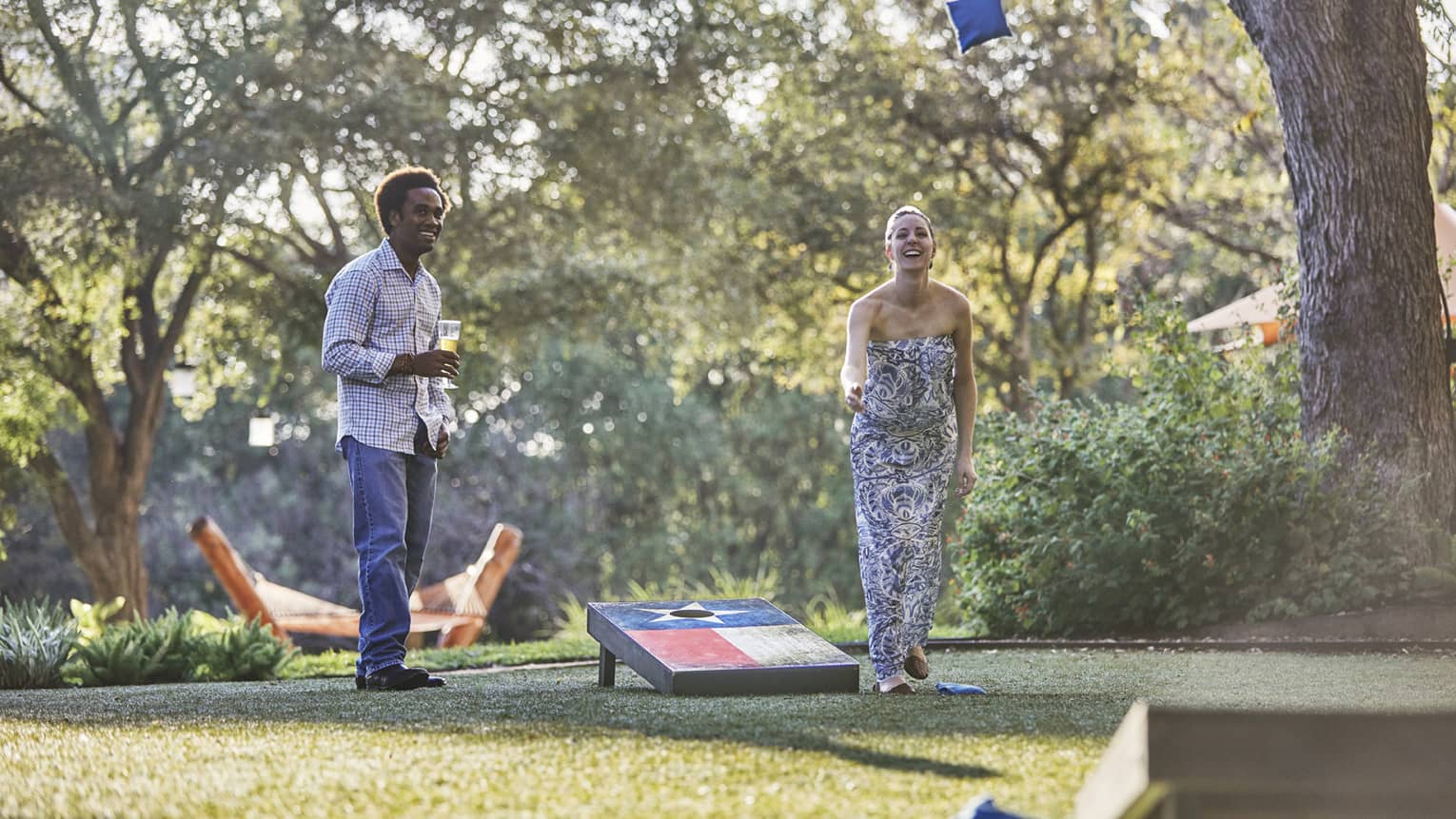 Laughing woman tosses bean bag on lawn, man holding beer watches