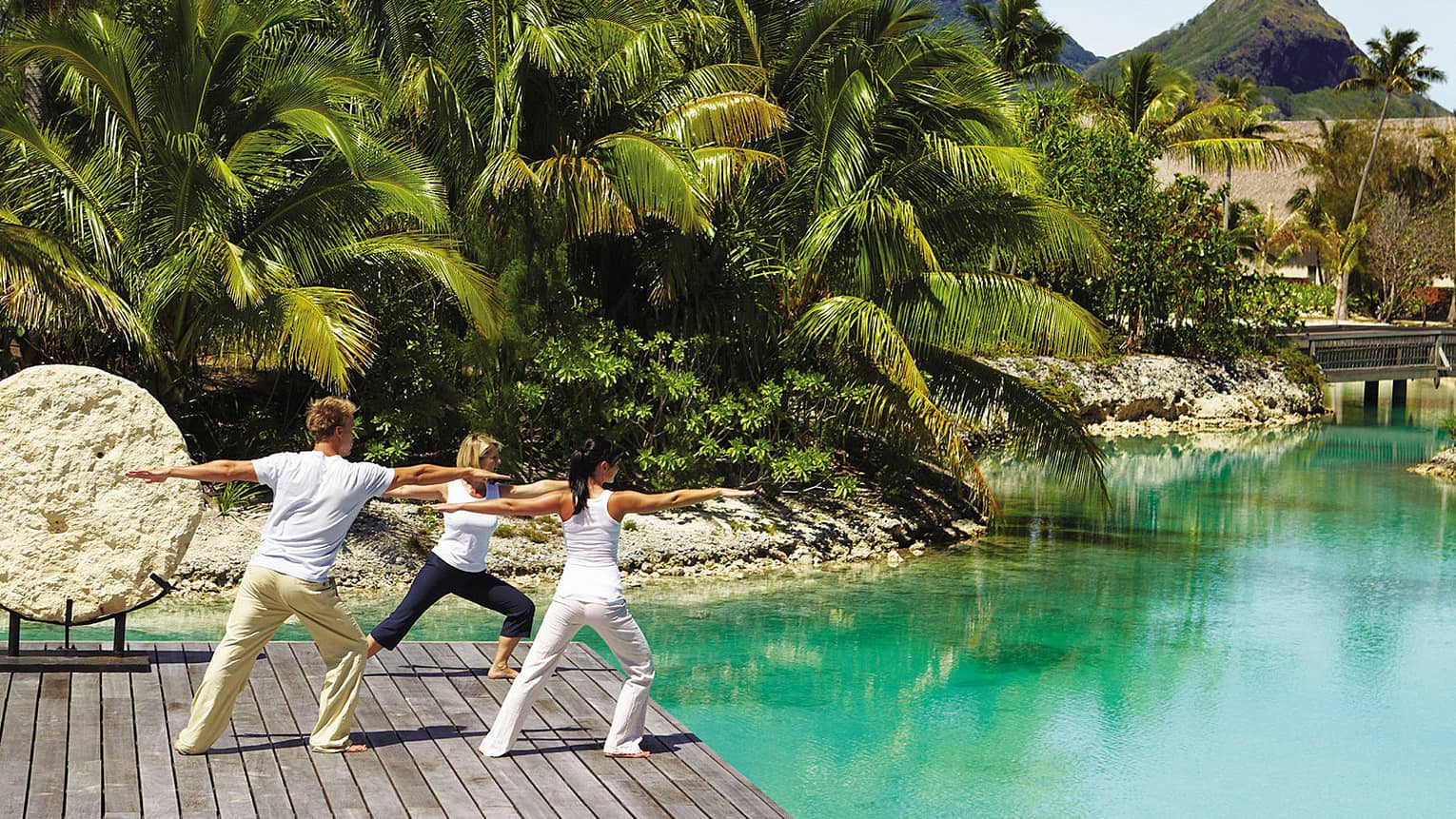 Man and two women stand on dock beside lagoon, with arms outstretched in yoga pose