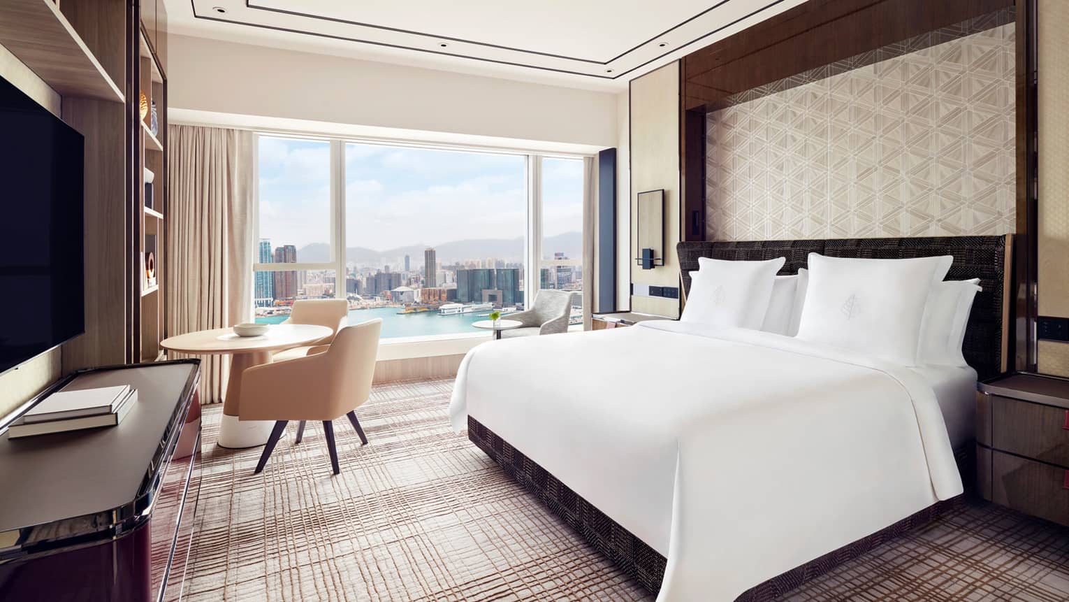 Modern hotel room with a large bed, and a round table by the window. Floor-to-ceiling windows offer a daytime view of the city skyline and waterfront.