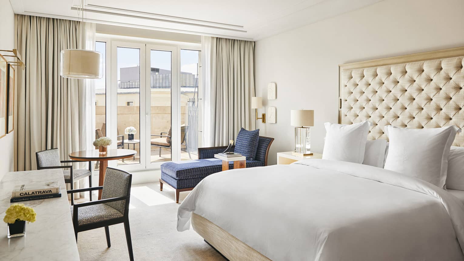 Luxury hotel bedroom with a king bed, tufted headboard, blue chaise lounge, round table and desk with chairs and French doors leading to a terrace