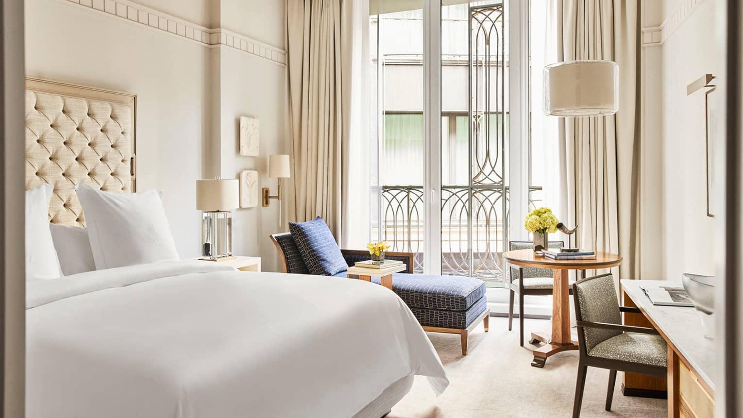 Luxury hotel bedroom with a king bed with tufted headboard, blue chaise lounge, round wood table with flowers and floor-to-ceiling windows with balcony view