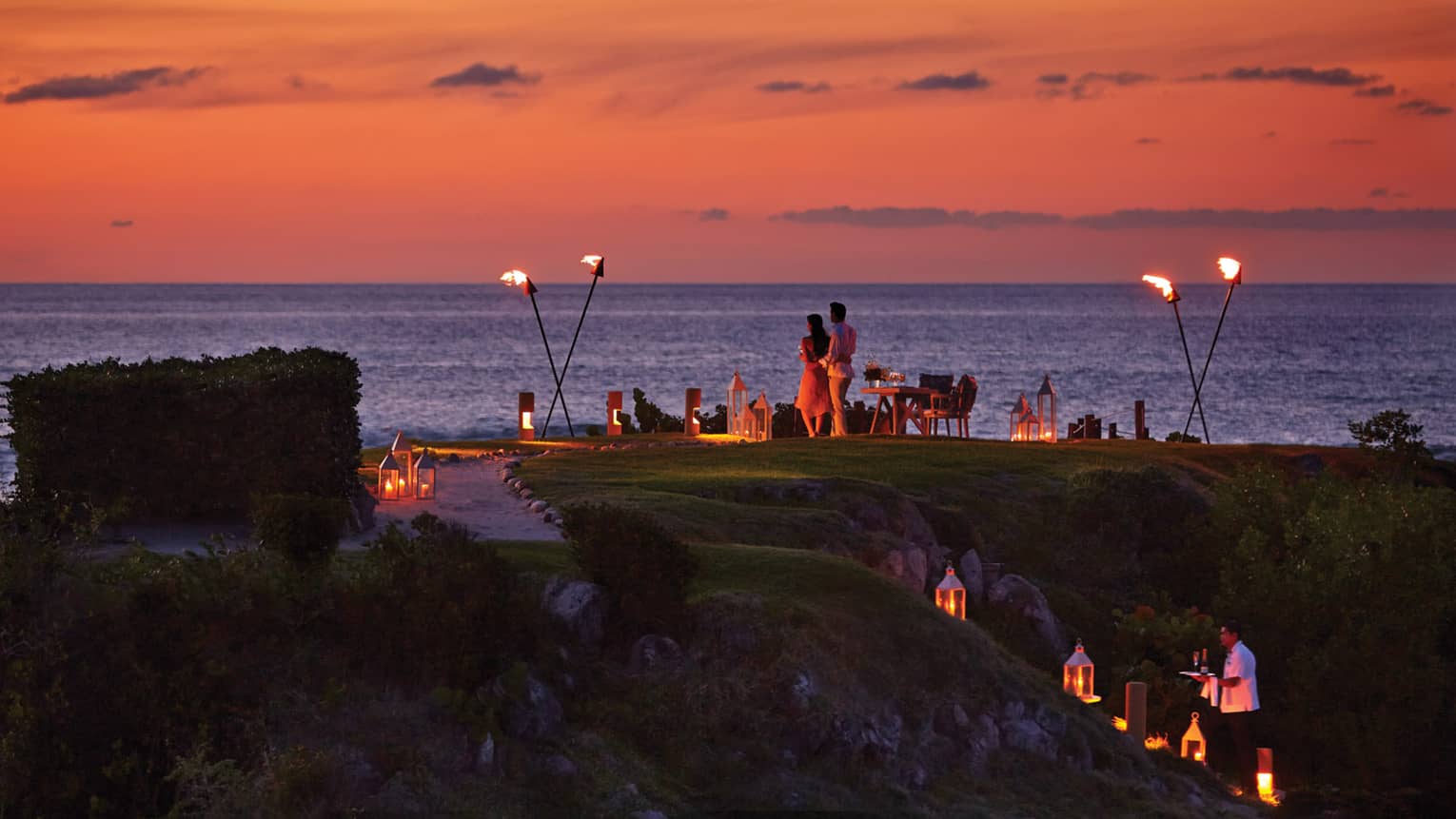 A couple stands on The Rock outpost at dusk, overlooking the ocean, with private dining table with lanterns and torches