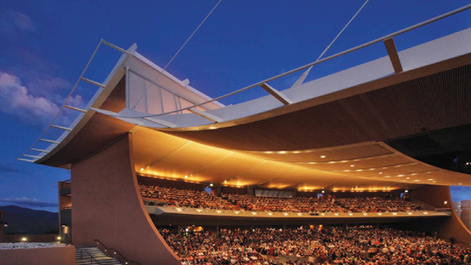 Curved opera building, audience in stands under lights at dusk