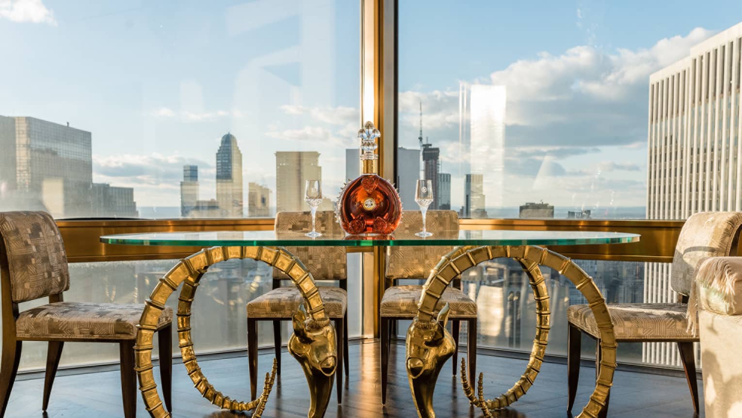 A bottle of LOUIS XIII Cognac and two glasses sit atop an ornate gold and glass dining table, in front of a floor-to-ceiling window overlooking New York City