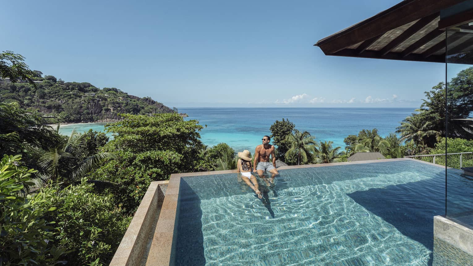 A couple lounges in a large infinity pool overlooking crystal blue waters