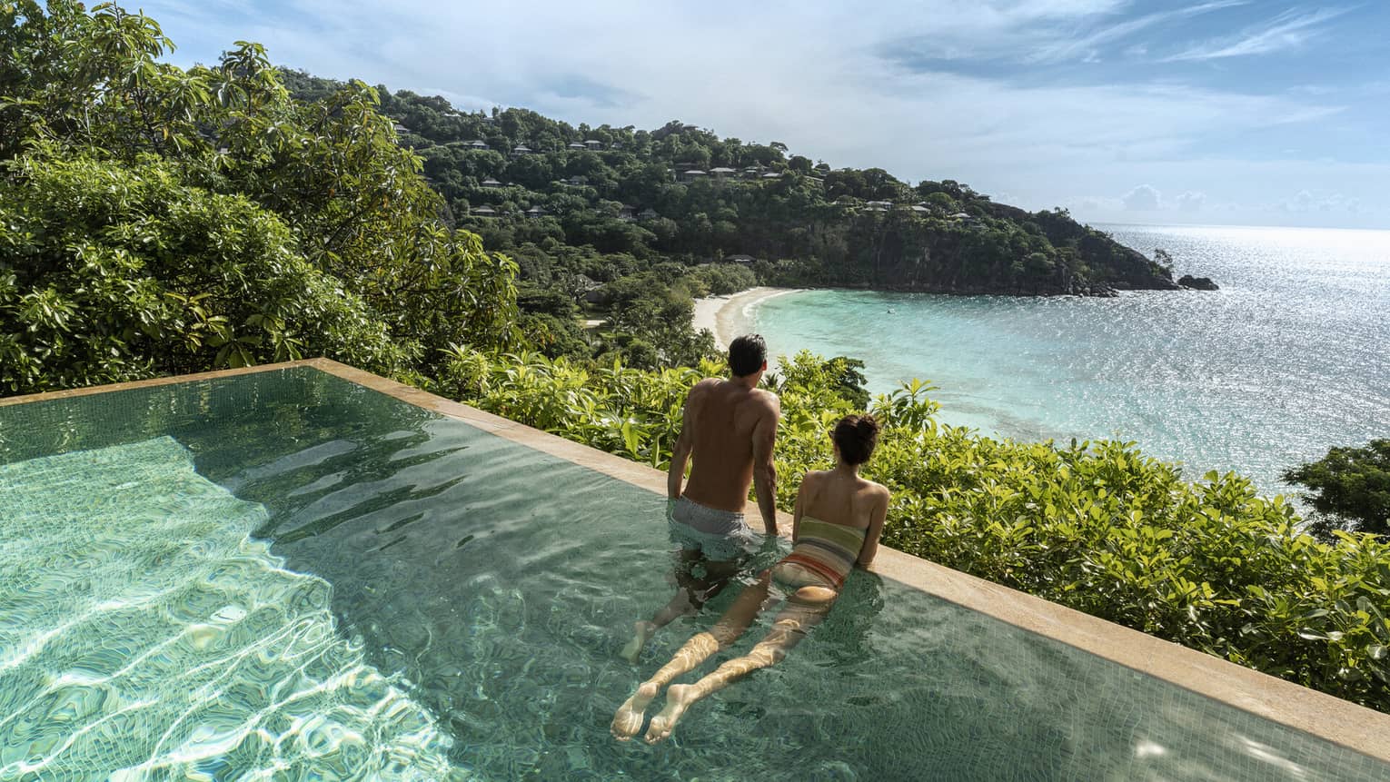 A couple looks out at a view of crystal blue waters and cliffs from an infinity pool