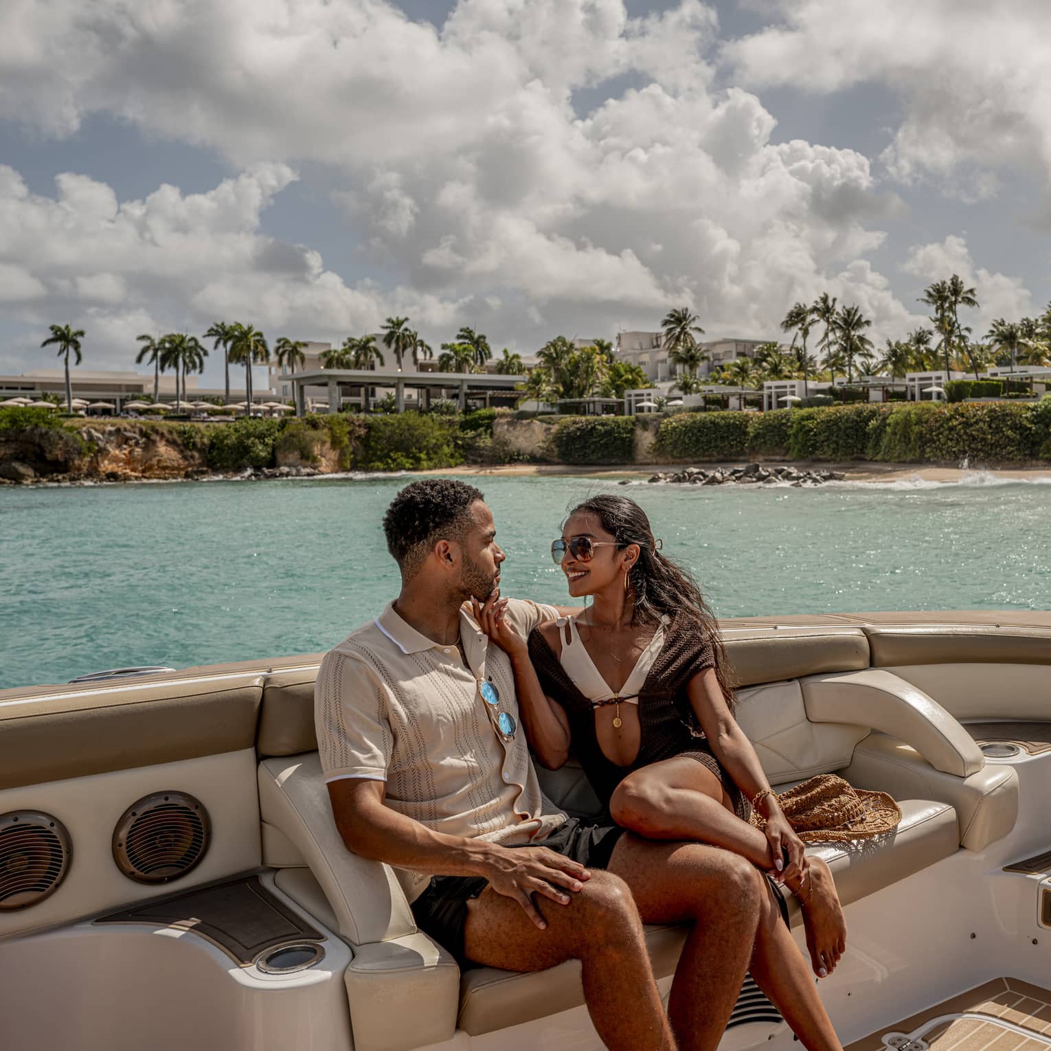 Two people sit on luxury yacht at sea, with palm trees and resort in the background