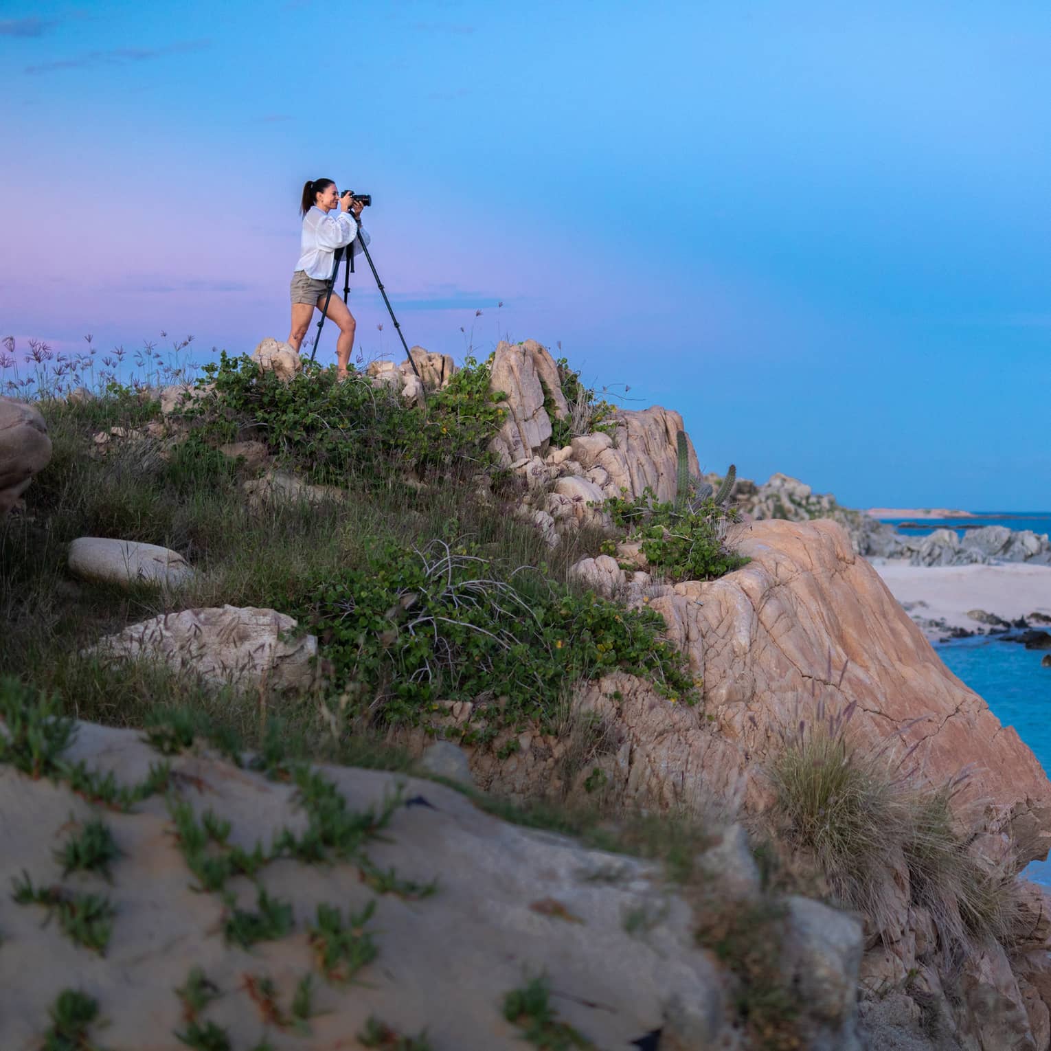 A photographer squints into a camera set on a tripod on a grassy rock outcrop overlooking calm, dark water under a full moon.