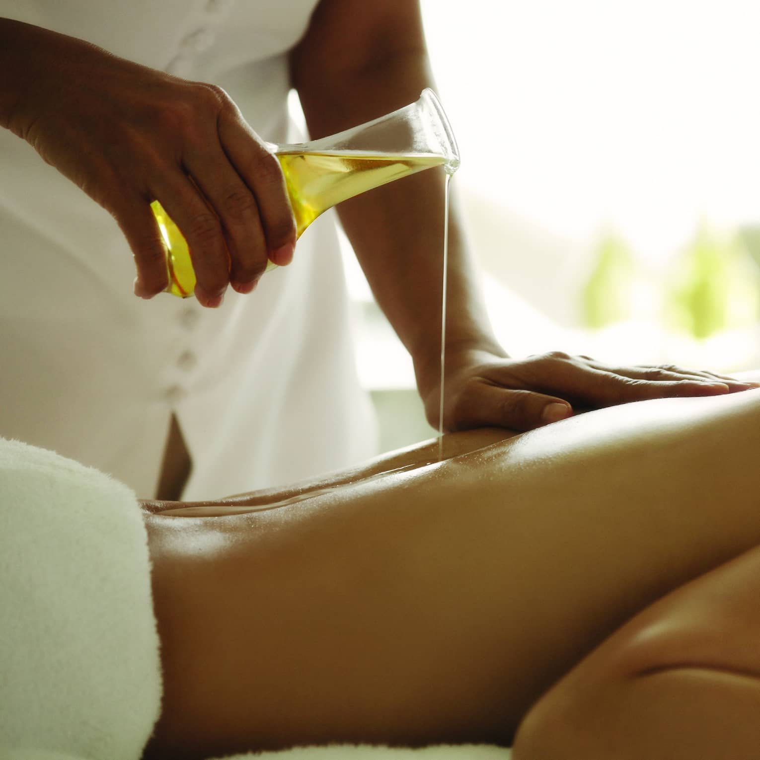 Spa staff pours massage oil over woman's bare back as she lays on treatment bed