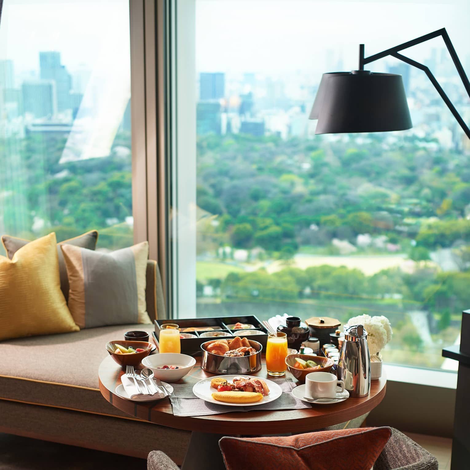 A breakfast tray of pastries, fresh fruit, juice and coffee on a guest room lounge chair at a window overlooking lush gardens.