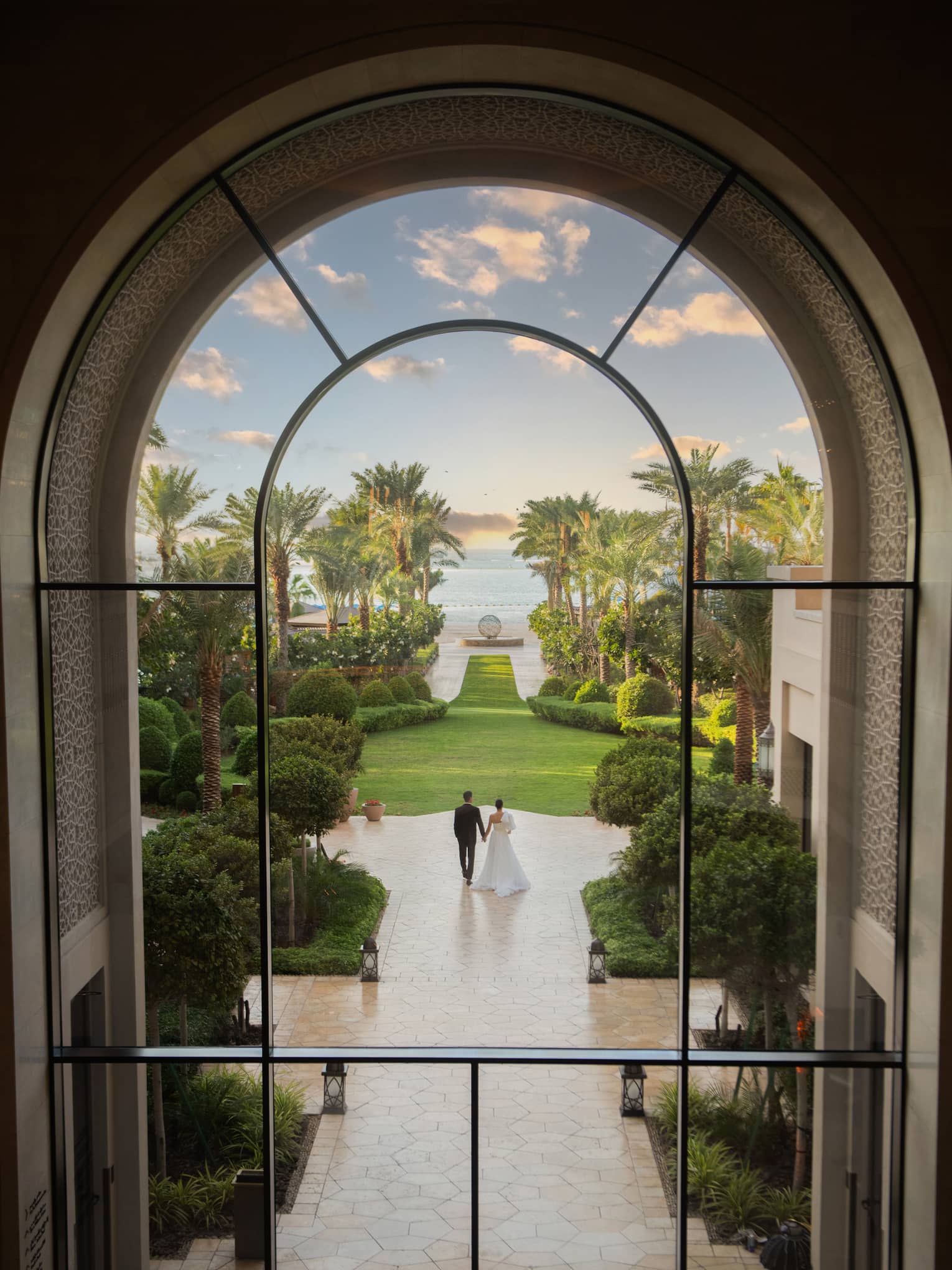 View through window looking out onto a bride and groom walk along a tiled outdoor pathway toward a green lawn with palm trees and the beach in the distance