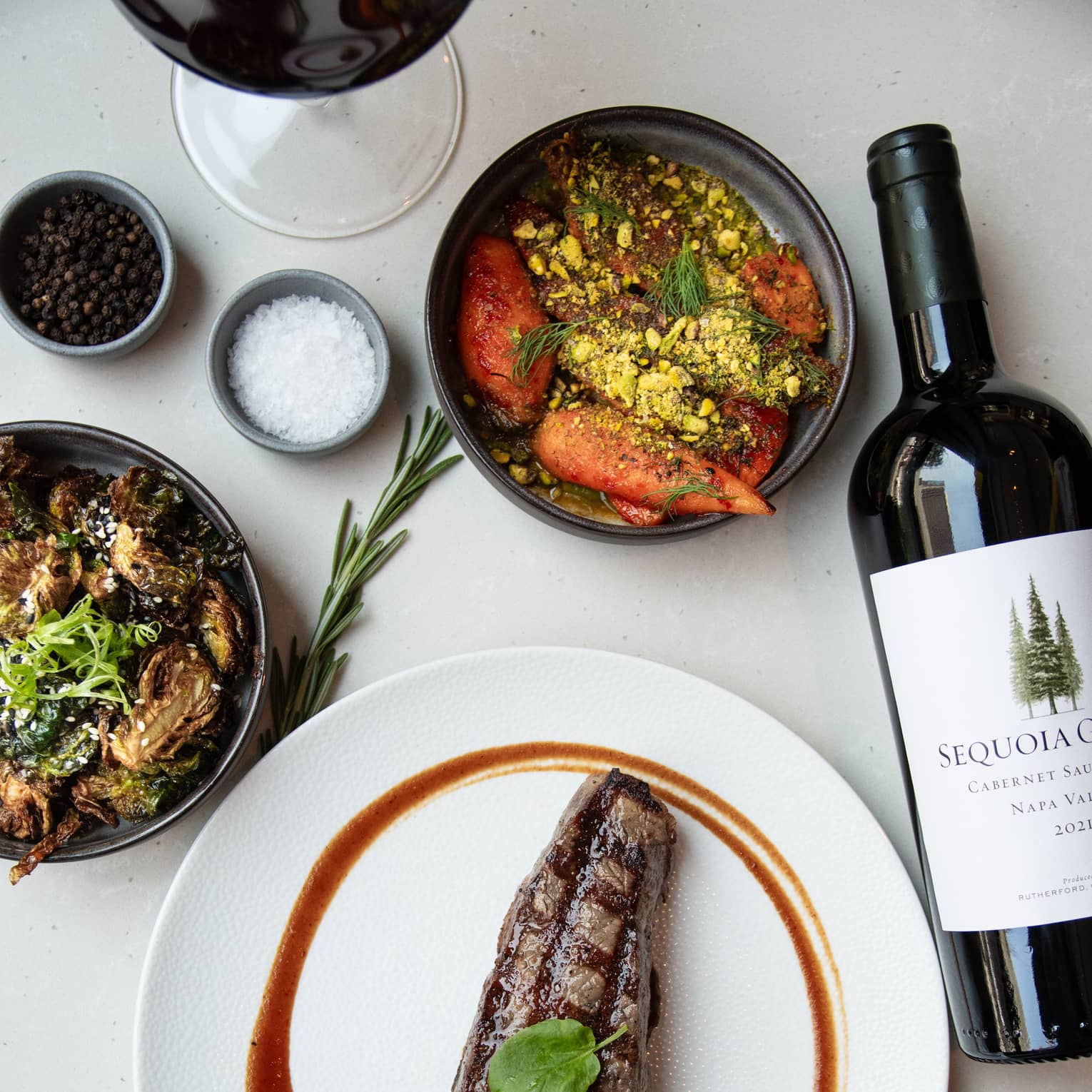 Strip steak on a white dish, bottle of wine, a small bowl of salt, small bowl of peppercorns, two bowls of food and a glass of wine on a white table