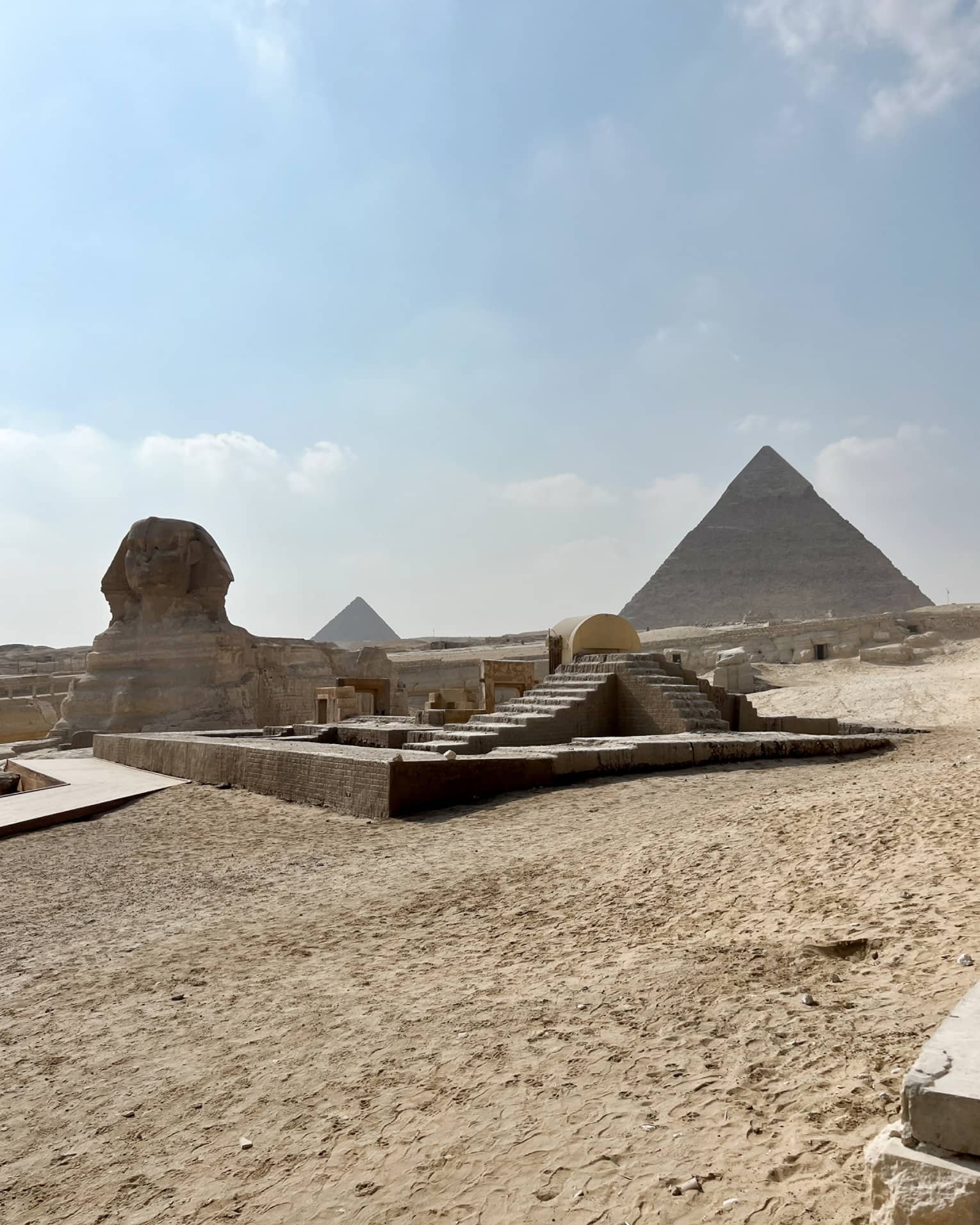 Distant view of the sphynx, desert sand and stone steps leading up to two pyramids under blue sky with fluffy clouds.