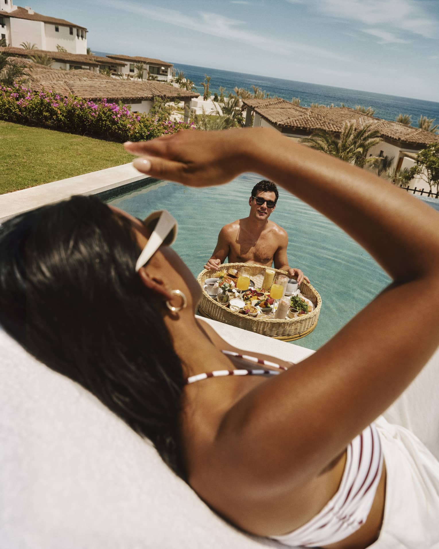 A guest stands in a pool with a floating basket of breakfast food and drinks and looks up at another guest sitting poolside.