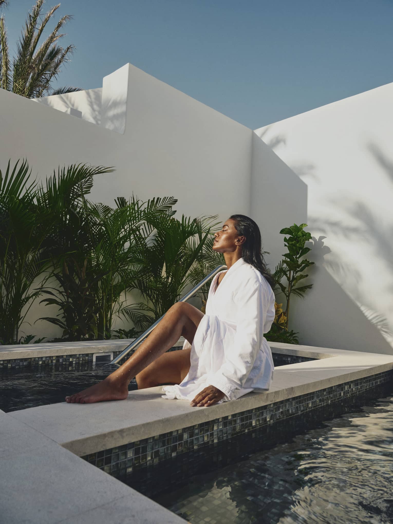 A guest in a linen bathrobe soaks in the sunlight while dipping their feet in a tiled pool surrounded by walls and plants.
