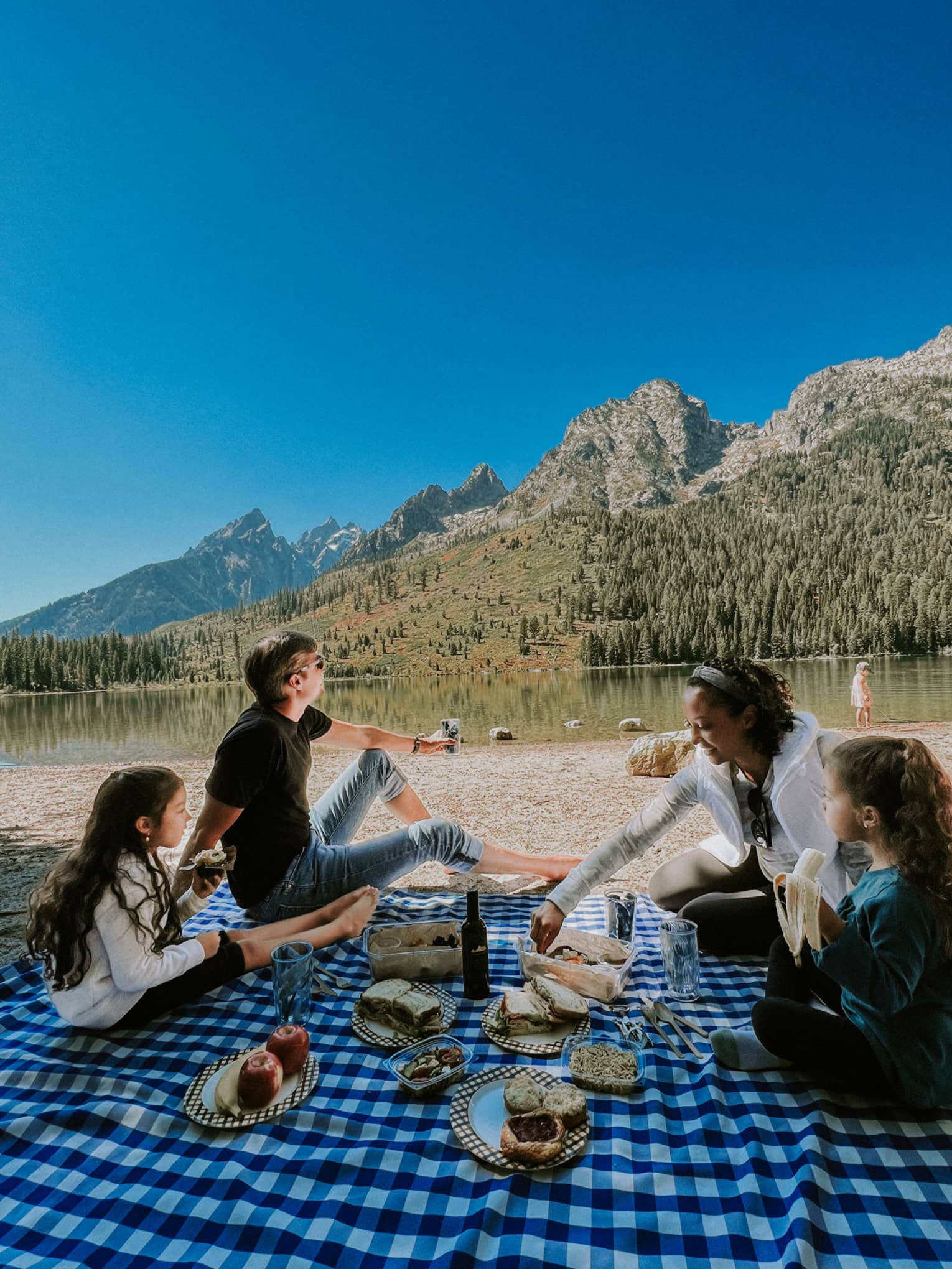 A family picnics on a checkered blanket near an alpine lake reflecting a rust and gold treed hill, distant mountains beyond,.