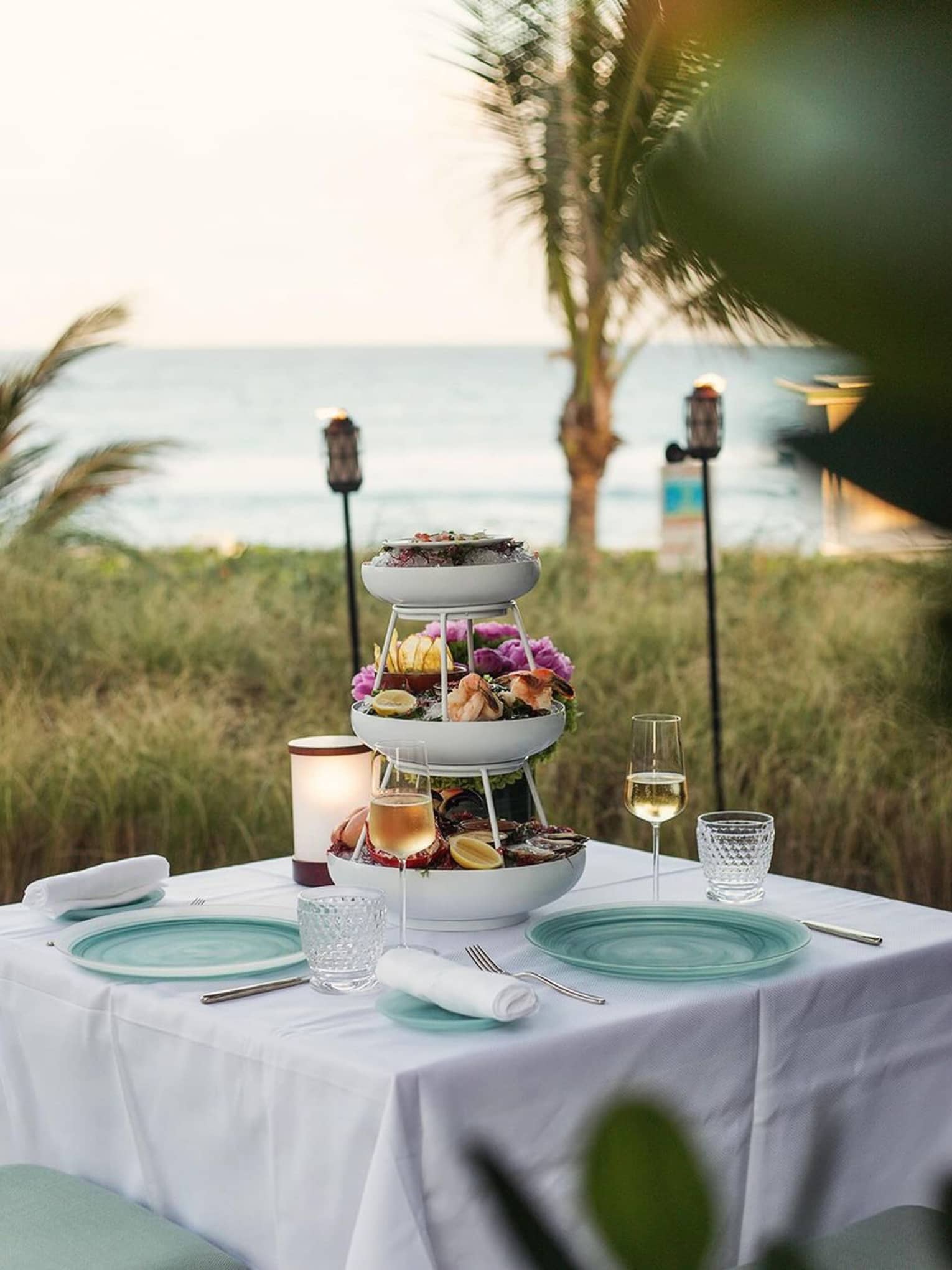 Three-tiered seafood tower is set in the centre of a dining table set outside with the beach nearby