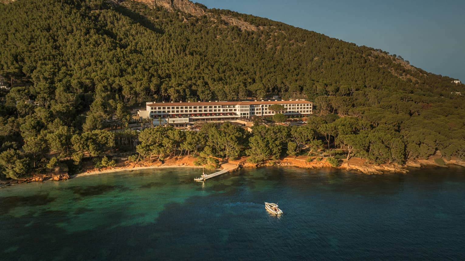 Late-day sunny aerial view of the bay and beach below the Four Seasons Resort nestled at the foot of a vast treed mountain.