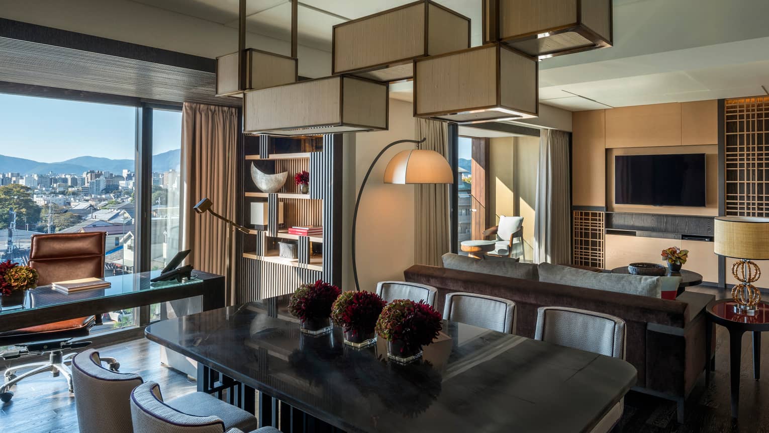 Residential suite with dining table, sofa, wall-mounted TV, floor lamp, bookshelf, desk with leather chair and floor-to-ceiling views of the city skyline