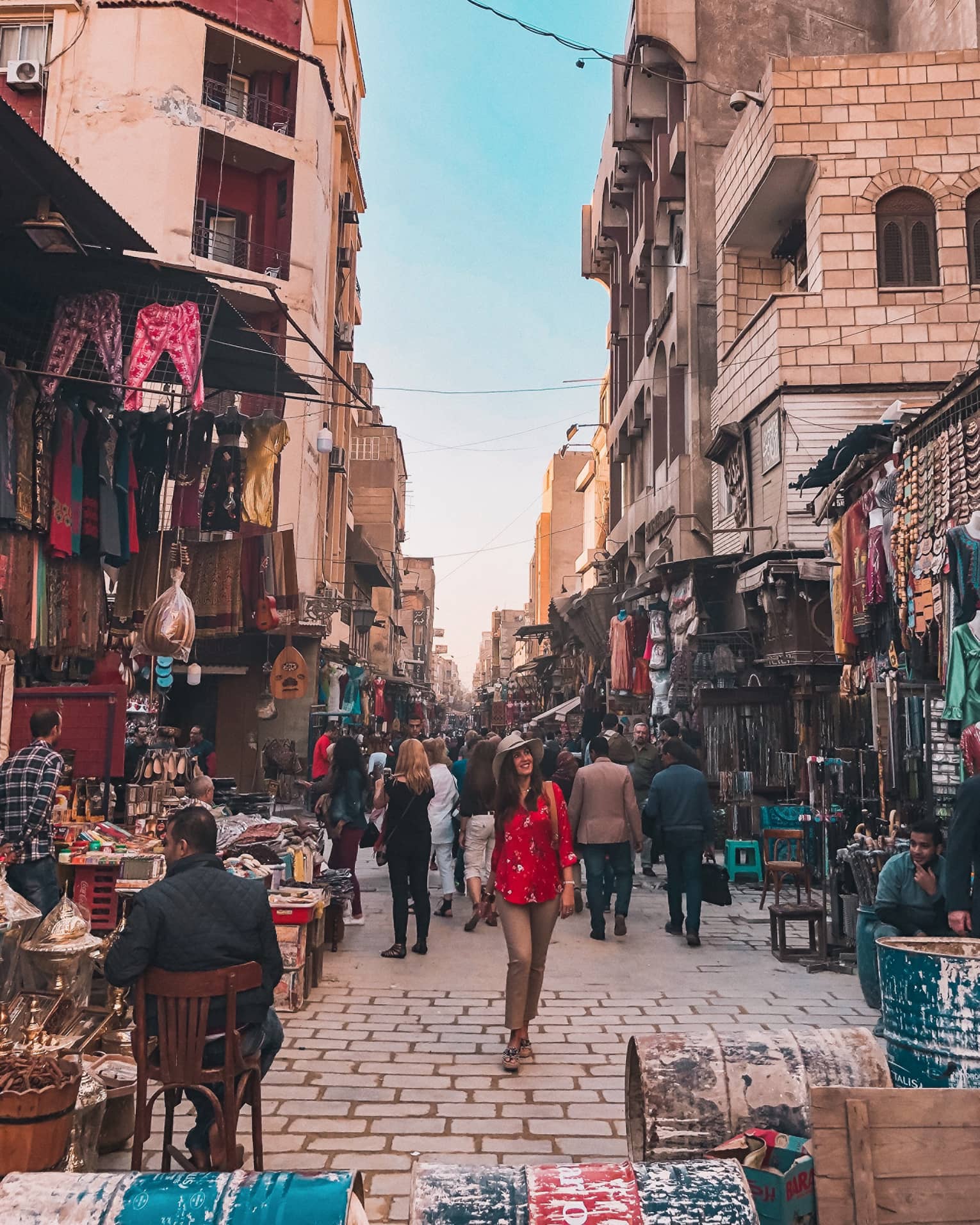 A smiling tourist strolls down a market alley, surrounded by varoius wares of the many merchants crowded between buildings.