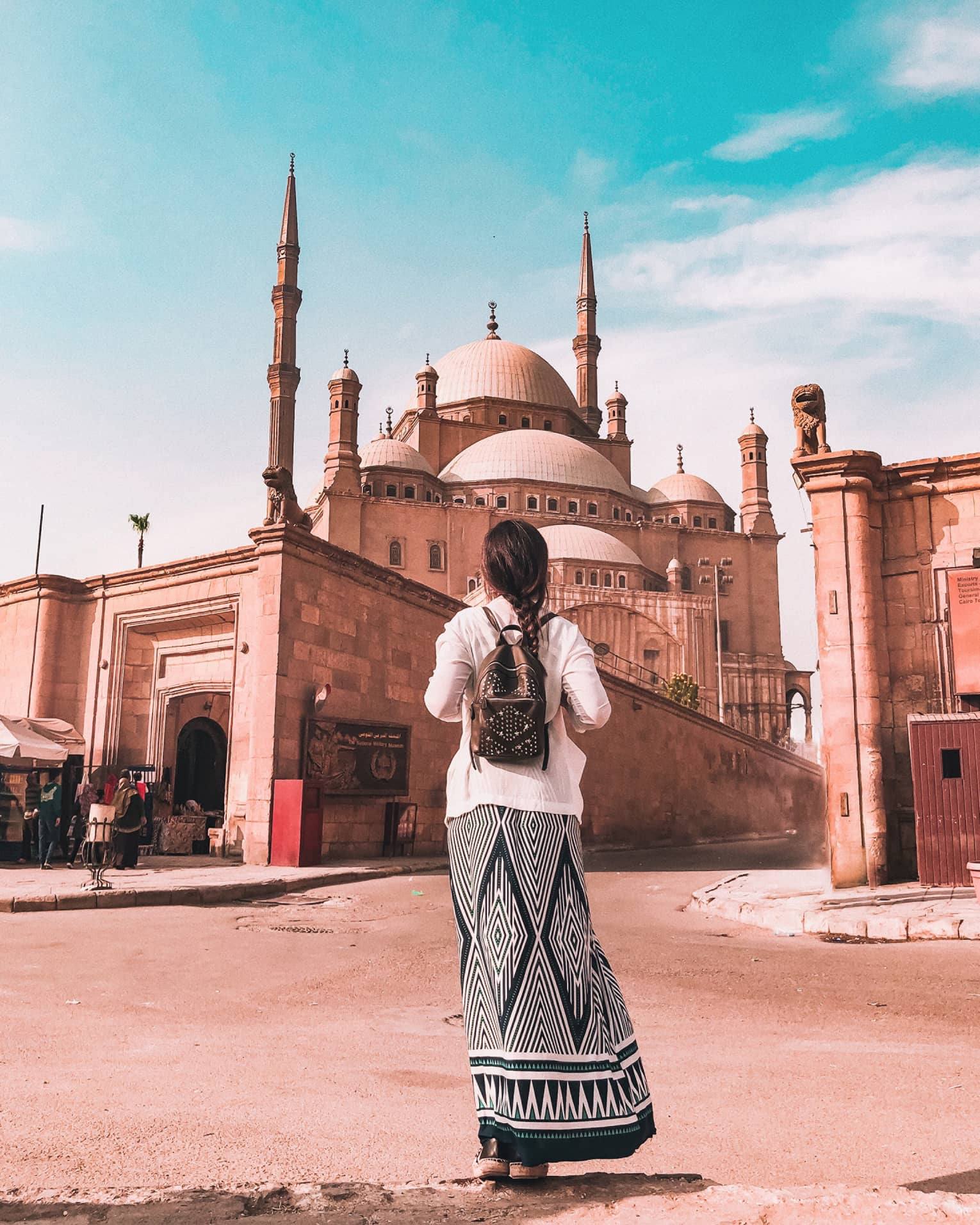 A tourist in a long skirt faces away, observing the Mosque of Muhammad Ali that towers over a wall surrounding it.