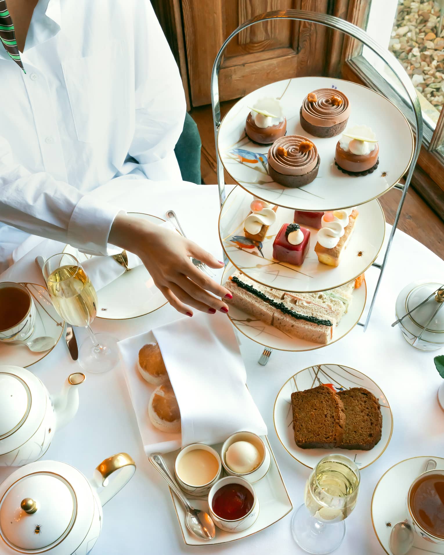 Person in white shirt reaches to classic afternoon tea stand filled with patisserie and mini sandwiches