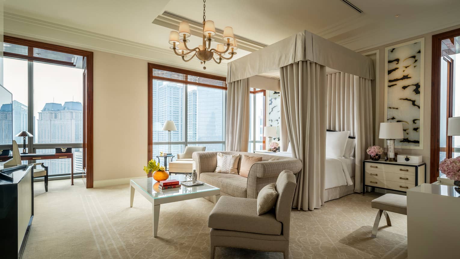 Hotel room canopy bed with white curtains behind sofa and seating area, large windows