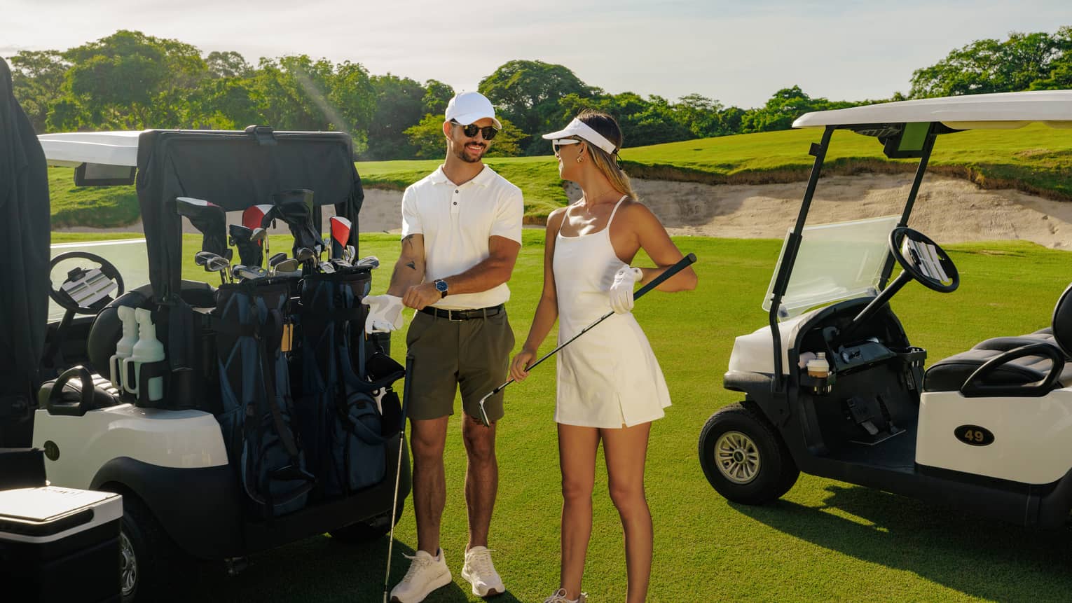 A man wearing a white polo shirt, green khaki shorts, white cap and sunglasses stands with a woman wearing a white tennis dress and a white visor in between two golf carts parked on a course