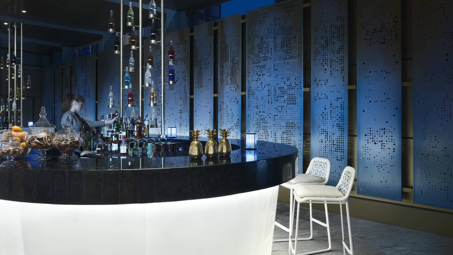 Bartender stands behind a round, black bar counter with white base, with two white stool placed nearby and colourful hanging bottles coming down from the ceiling in the centre of the bar