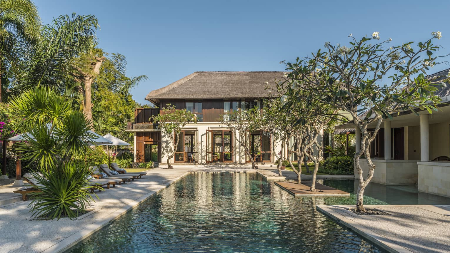Two-storey villa at end of long outdoor swimming pool lined with trees, tropical plants