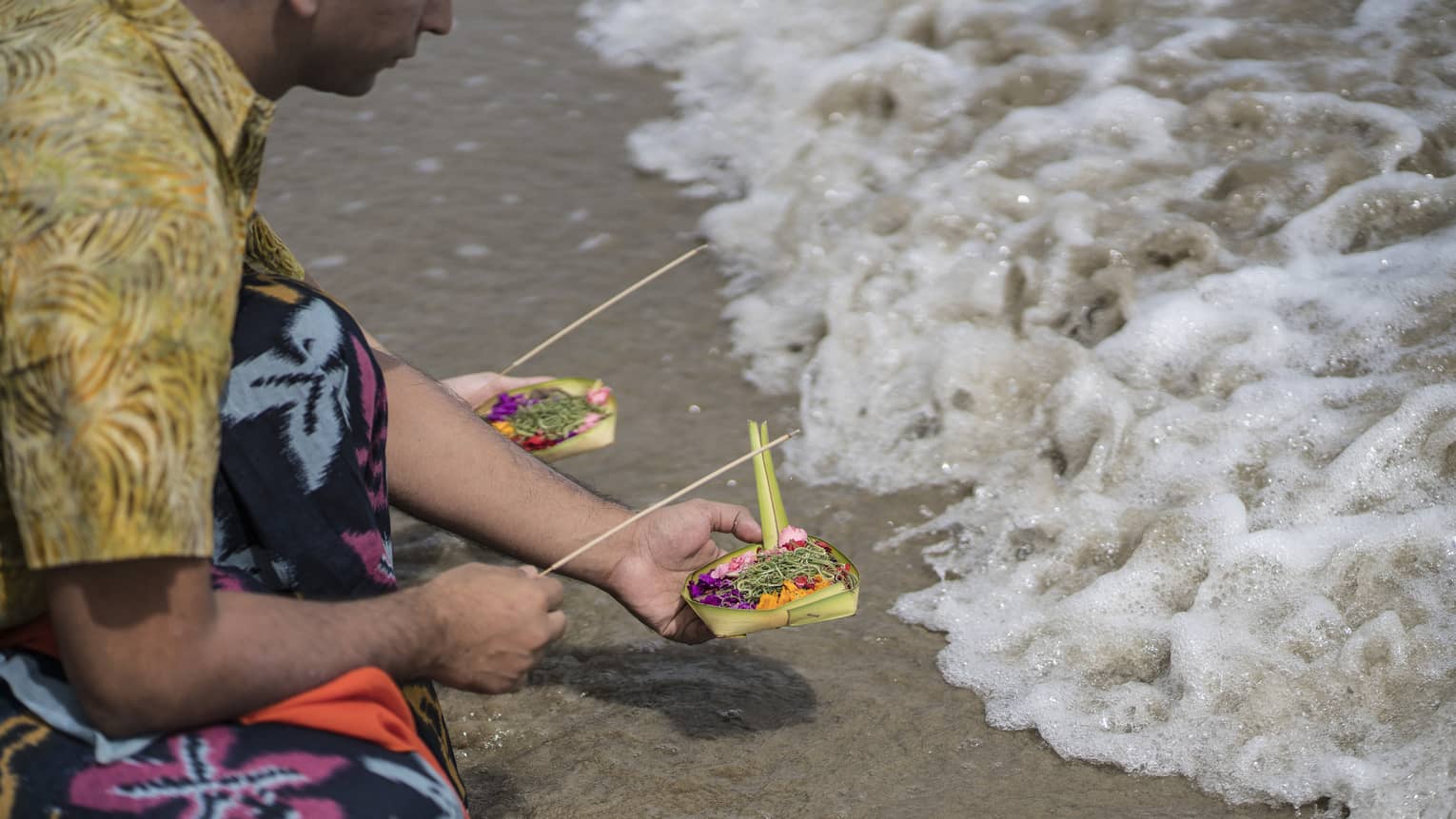 Kneeling at the edge of frothy water, a person in colourful ceremonial dress offers incense and a small woven box of flowers.