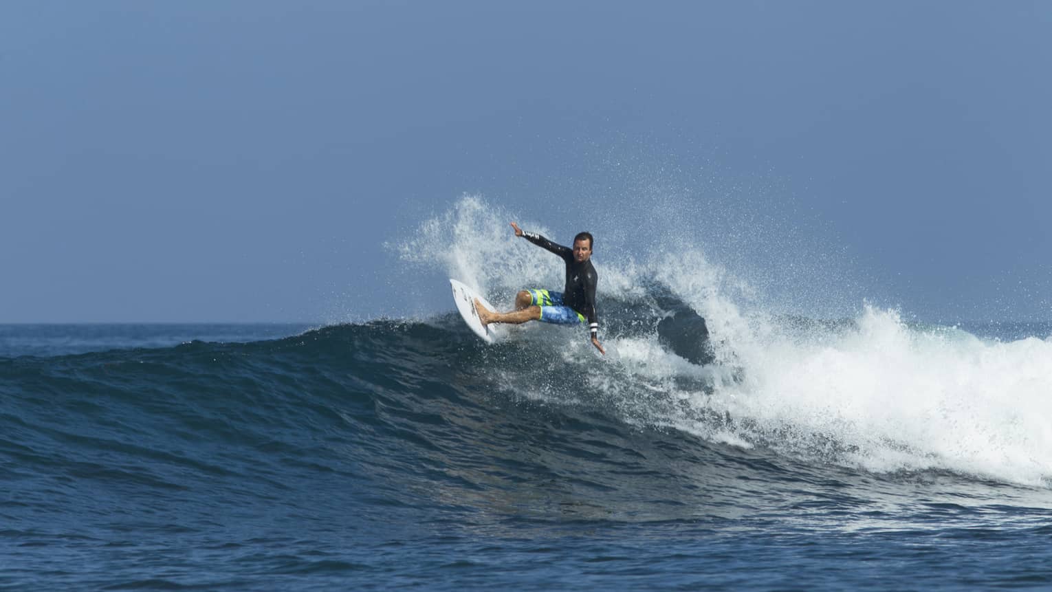 A surfer in a long sleeve rash guard and board shorts rides a wave, arms outstretched, in the wake of frothy white spray.