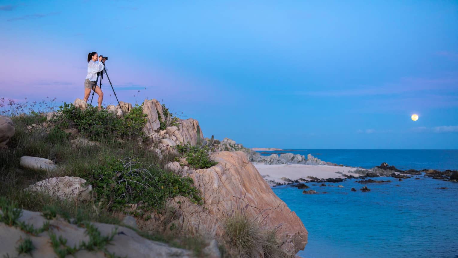 A photographer squints into a camera set on a tripod on a grassy rock outcrop overlooking calm, dark water under a full moon.