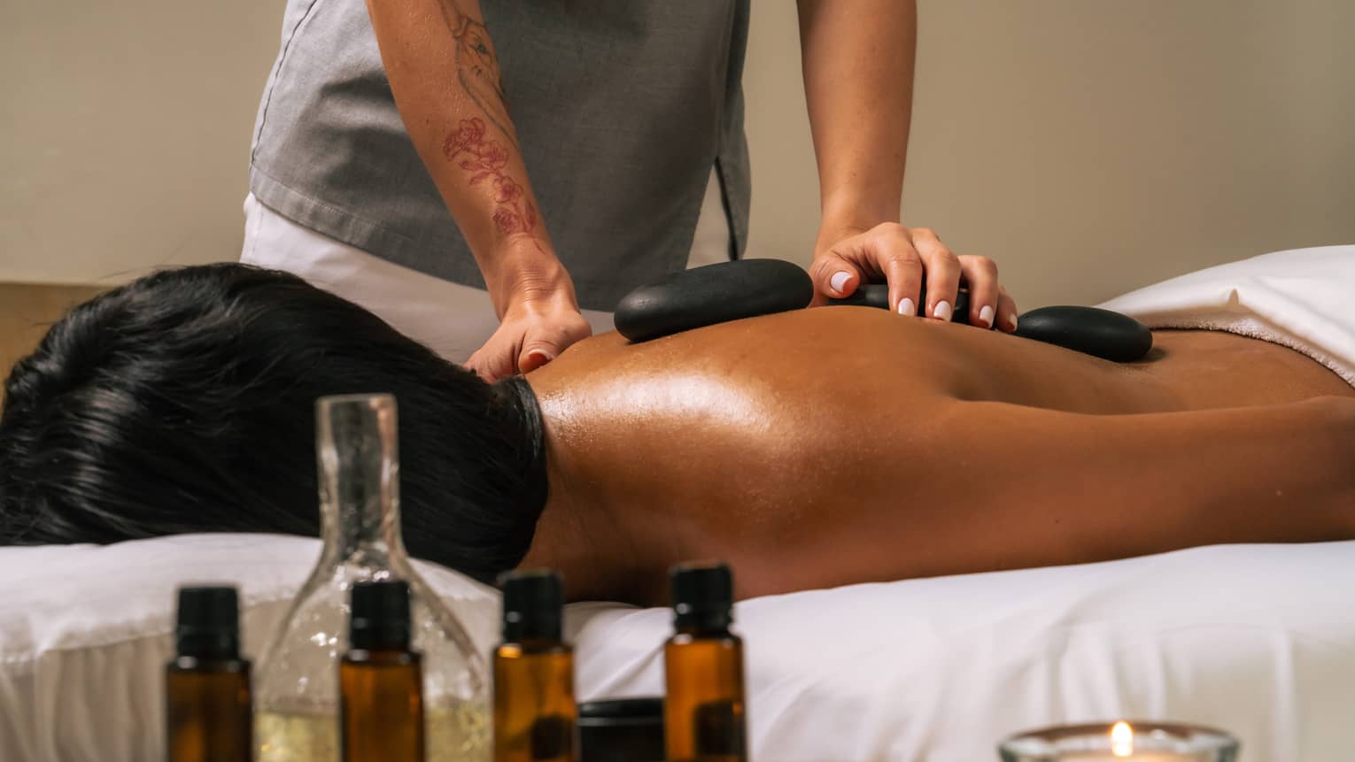 Person lies face down as a massage therapist places black stones on their bare back, with various bottles of oil in the foreground