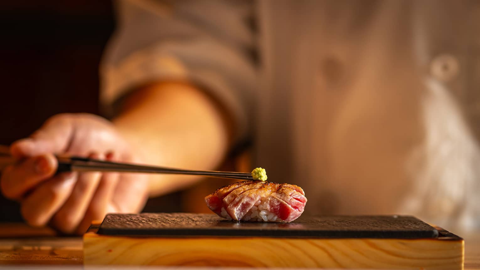 Seared tuna nigiri topped with a small dab of wasabi, placed on a black slate plate with a warm, blurred background