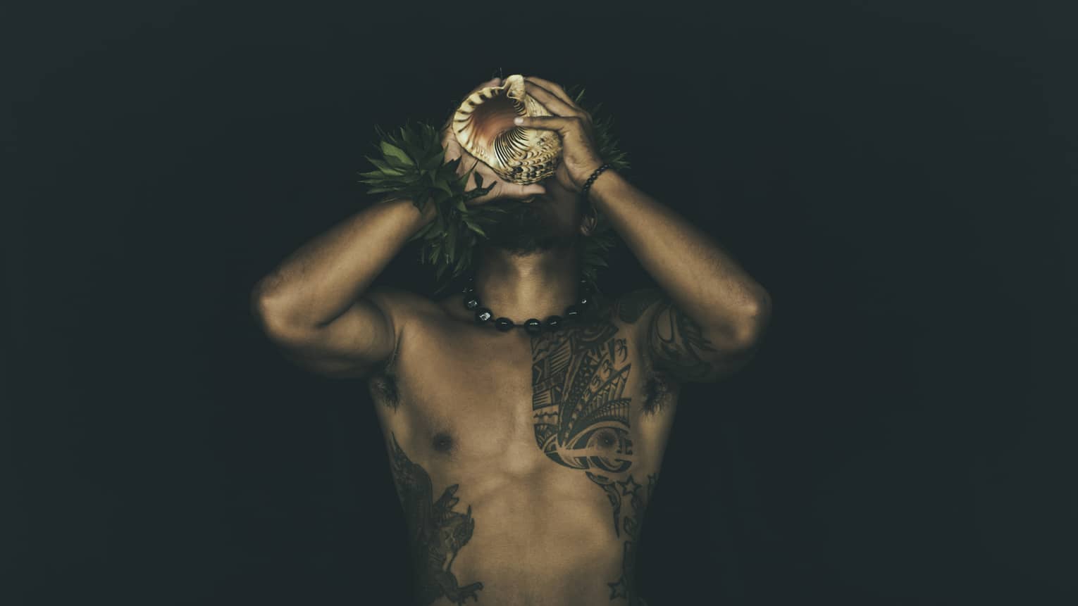 Shirtless tattooed Kānaka Maoli man plays traditional conch shell like a trumpet in the night
