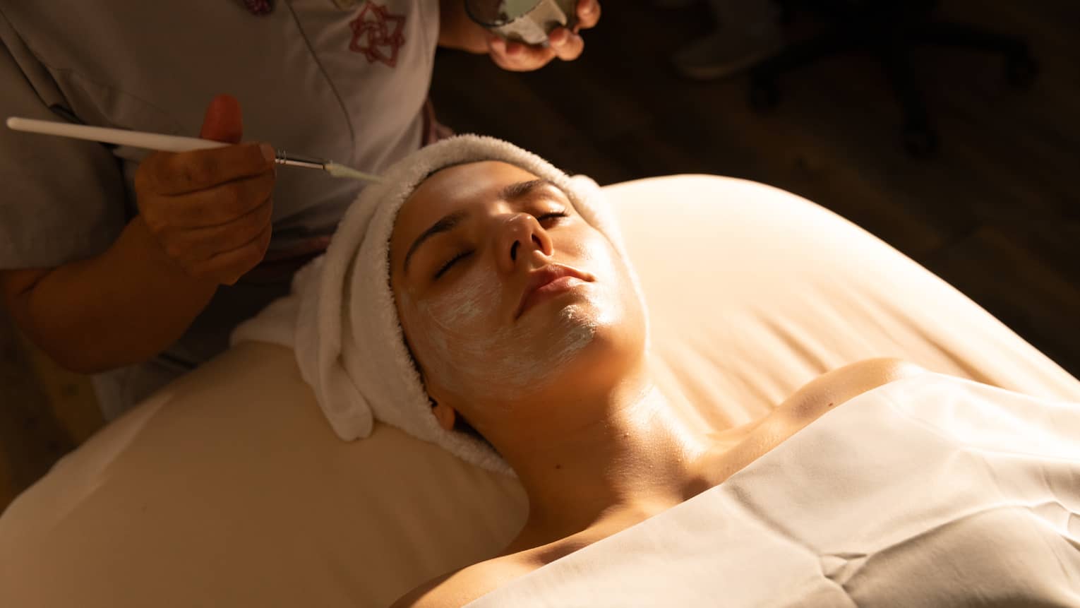 A person with draping over their body and a towel wrap on their head lies on a spa treatment table and receives a facial treatment.