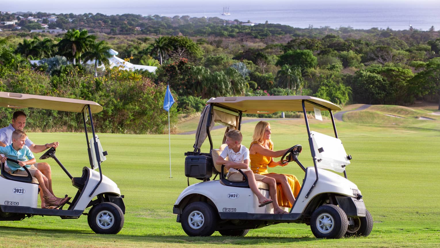Two adults driving golf carts past a golf green, each with a smiling child passenger, trees and ocean visible beyond.