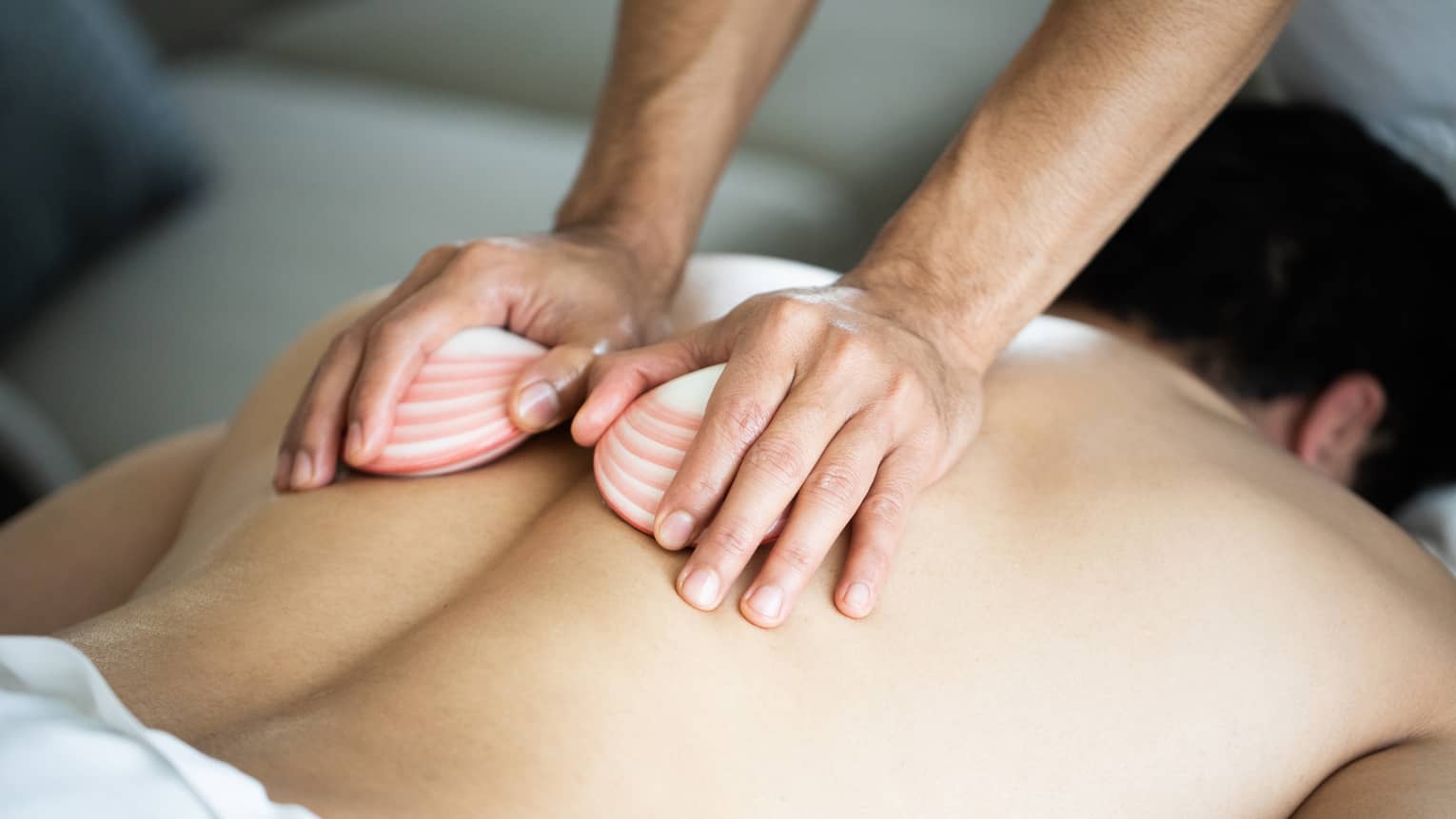 A masseuse's hands hold two white, pink-striped lava shells, running over the bare back of a man lying face down on a massage table