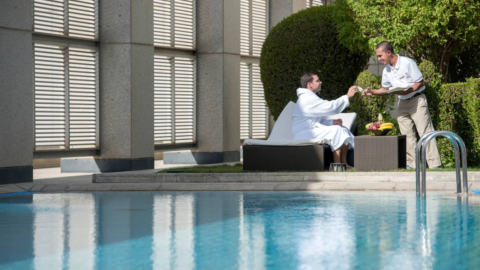 Man in white bathrobe sits on lounge chair on sunny swimming pool deck by fruit bowl, hotel stands hands him cloth