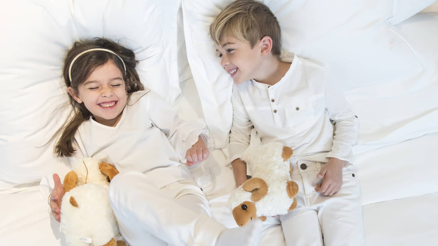 Aerial view of two children wearing white pyjamas with white stuffed animals lying on bed, laughing