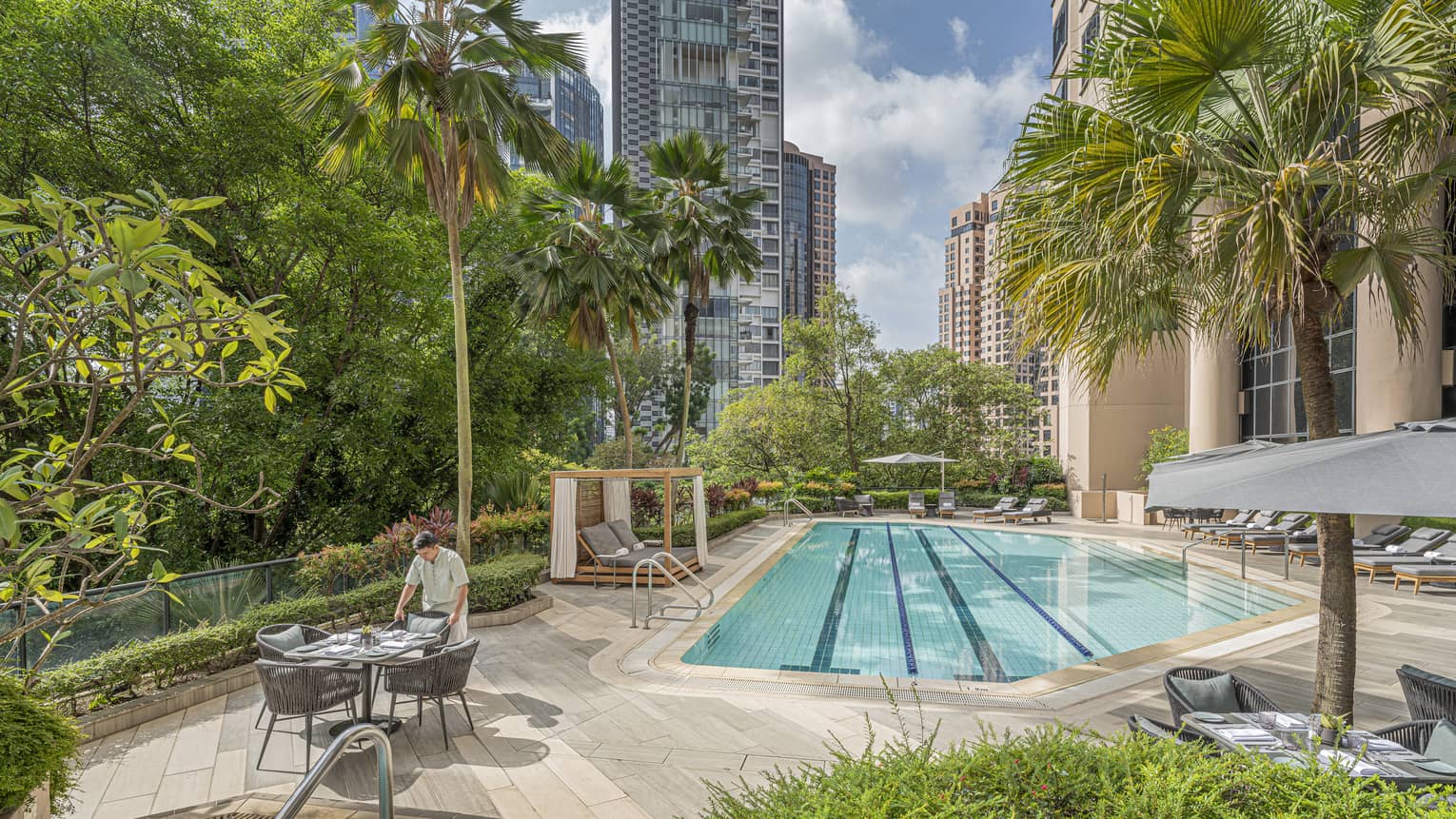 Outdoor pool area of a hotel, surrounded by lush greenery and tall buildings, with a staff member setting up a table beside a jetted tub and chaise lounges