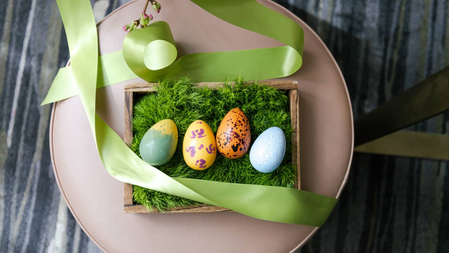 Four multi-colored chocolate Easter eggs on a bed of green moss set in a wooden box with a light-green ribbon draped around it