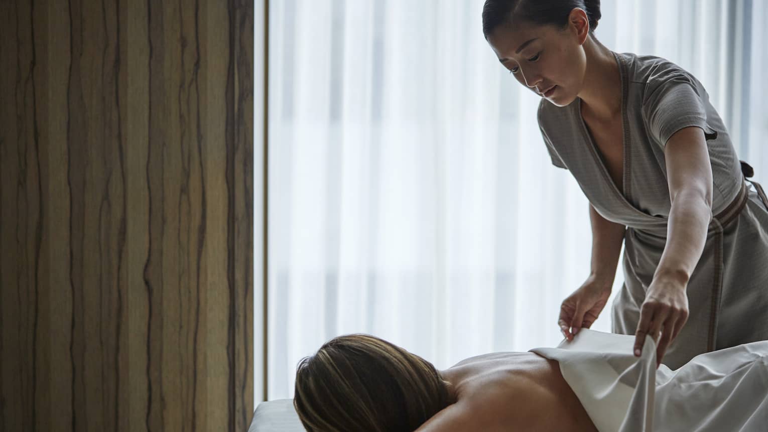 A masseuse covers a woman's back with a sheet during a massage.