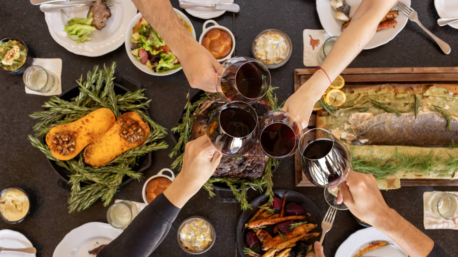 Overhead view of a table filled with festive dishes such as roasted squash, a filet of fish and other platters with four hands each holding a glass of red wine coming together in the centre