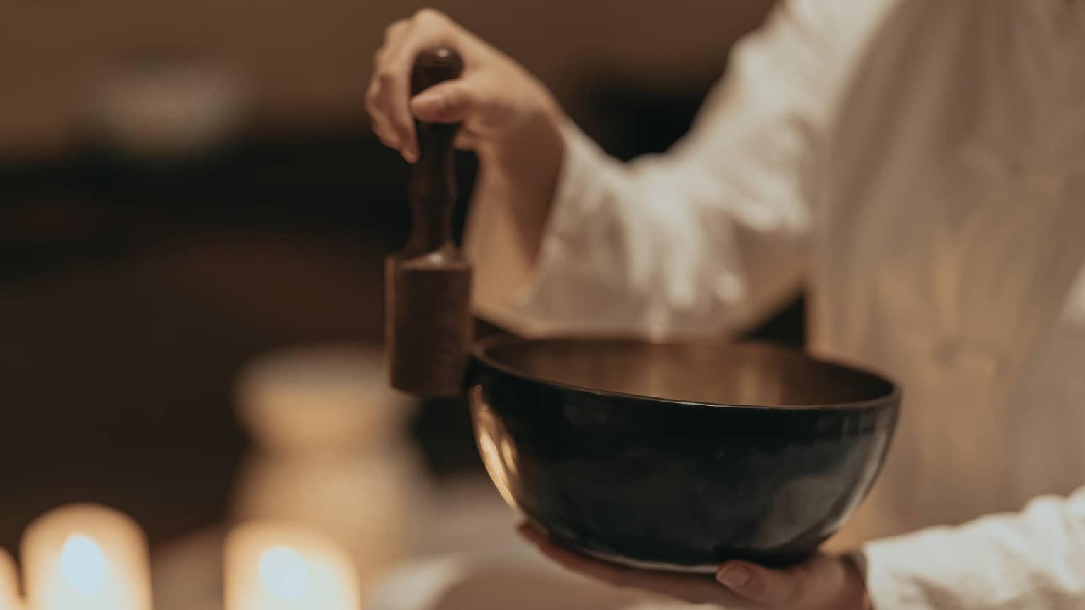 Close-up of an instructor rubbing a mallet against the edge of a sound bowl in a dimly lit room surround by burning candles.